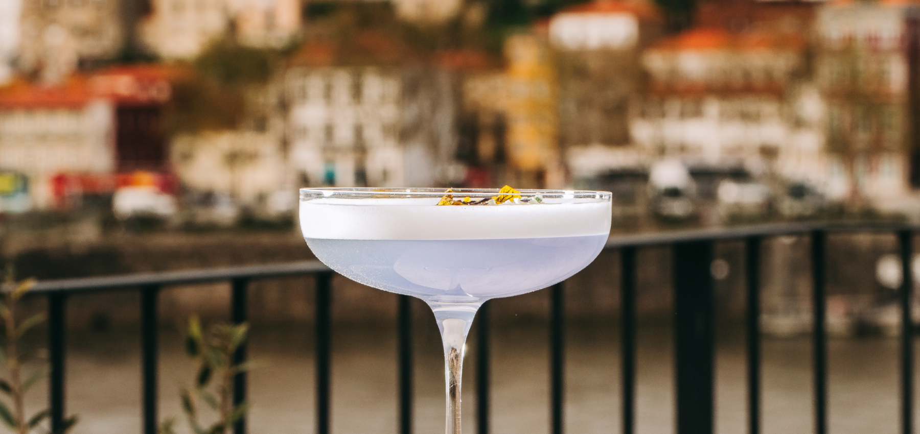 A close-up of a cocktail with a pale purple hue in a coupe glass, garnished with dried flowers, set against a blurred cityscape and river background.