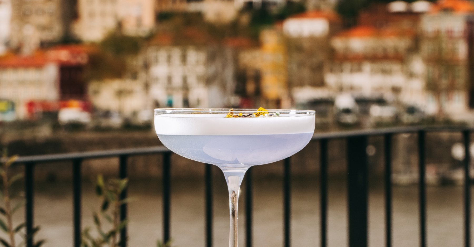 A close-up of a cocktail with a pale purple hue in a coupe glass, garnished with dried flowers, set against a blurred cityscape and river background.