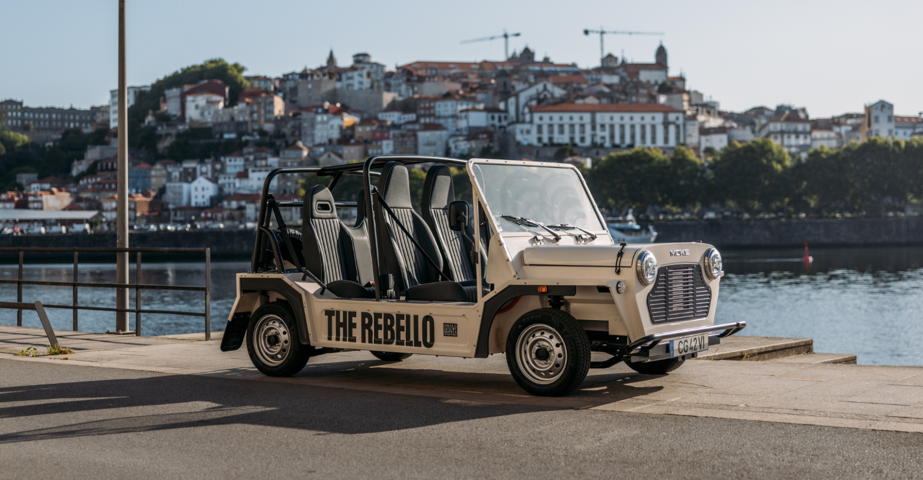 A cream-coloured open-air vehicle labelled "THE REBELLO" is parked by a riverside. In the background, there is a scenic view of a city with hillside buildings and cranes.