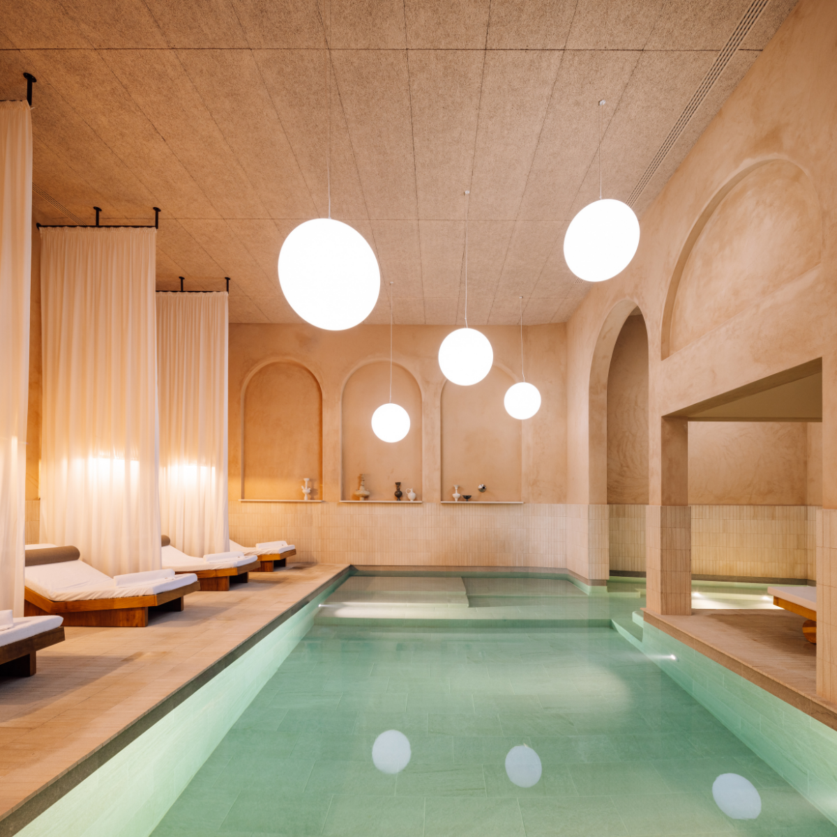 Modern indoor spa at Hotel Porto, featuring a turquoise pool, beige walls, arched alcoves, glowing round lights, and cushioned loungers separated by white curtains. The atmosphere is calm and inviting.