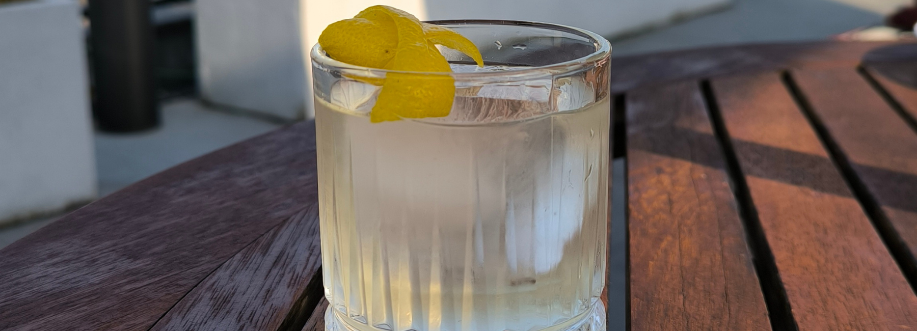 A clear cocktail with ice and a lemon twist garnish sits on a wooden outdoor table in sunlight.