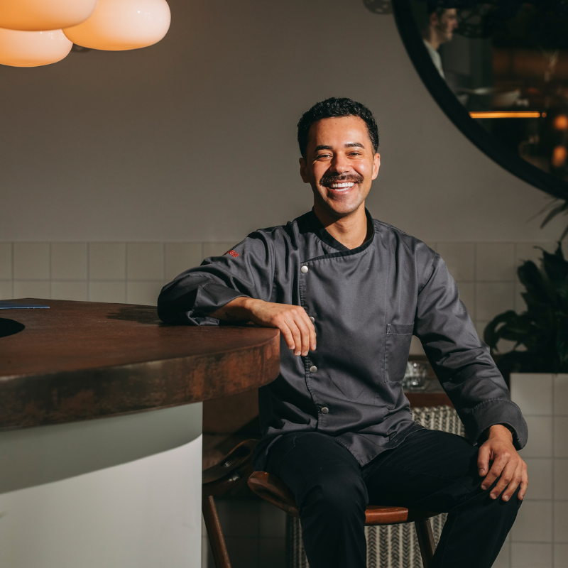 A smiling restaurant chef in a dark uniform sits on a chair by a curved worktop in a warmly lit modern kitchen, with plants and a round mirror in the background.