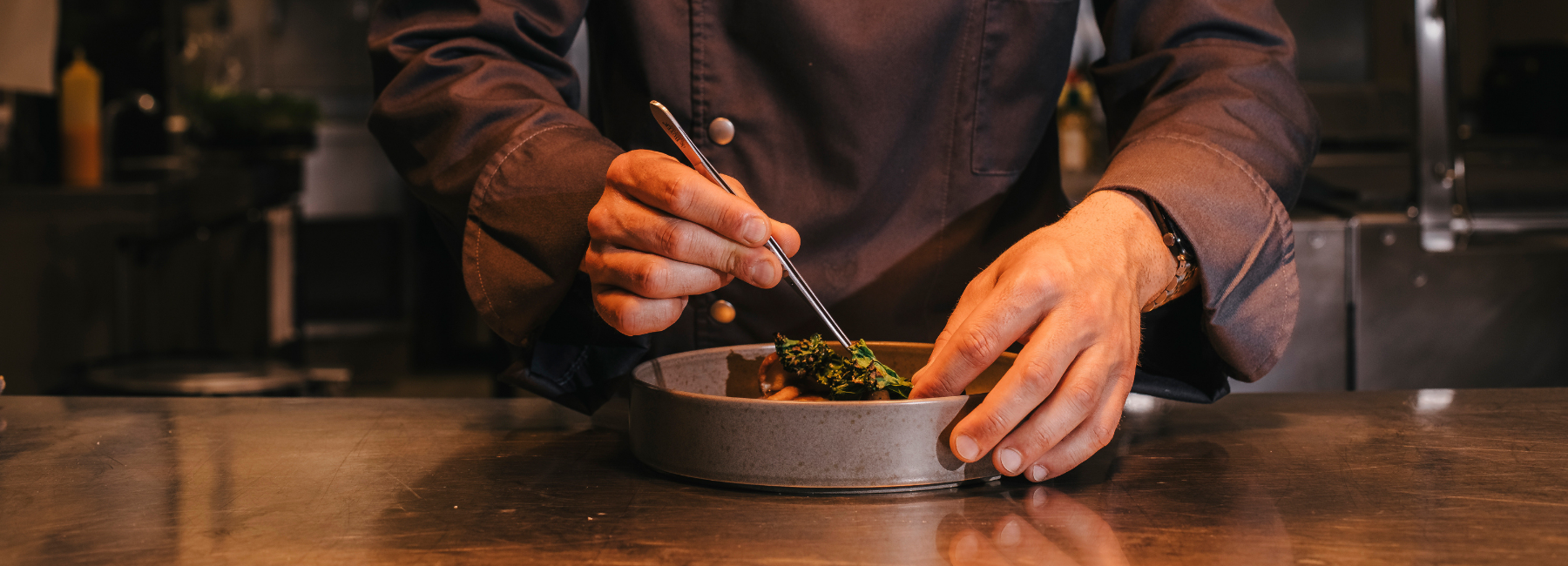A restaurant chef in a dark uniform uses tweezers to carefully plate food in a kitchen, focusing on precise presentation of greens on a dish.