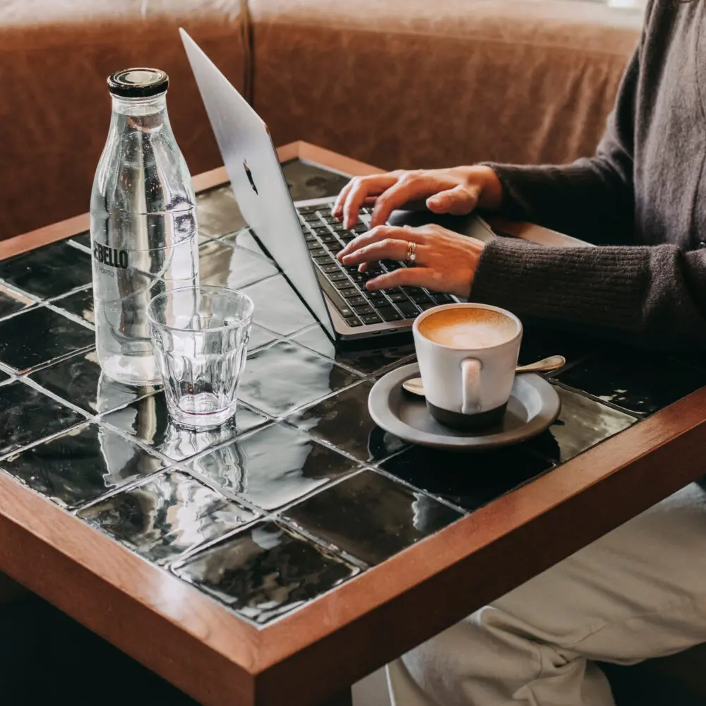 A person types on a laptop at a tiled café table with a cup of coffee, a glass, and a glass water bottle, with the windows revealing the outline of a nearby hotel building. Only their hands and torso are visible.