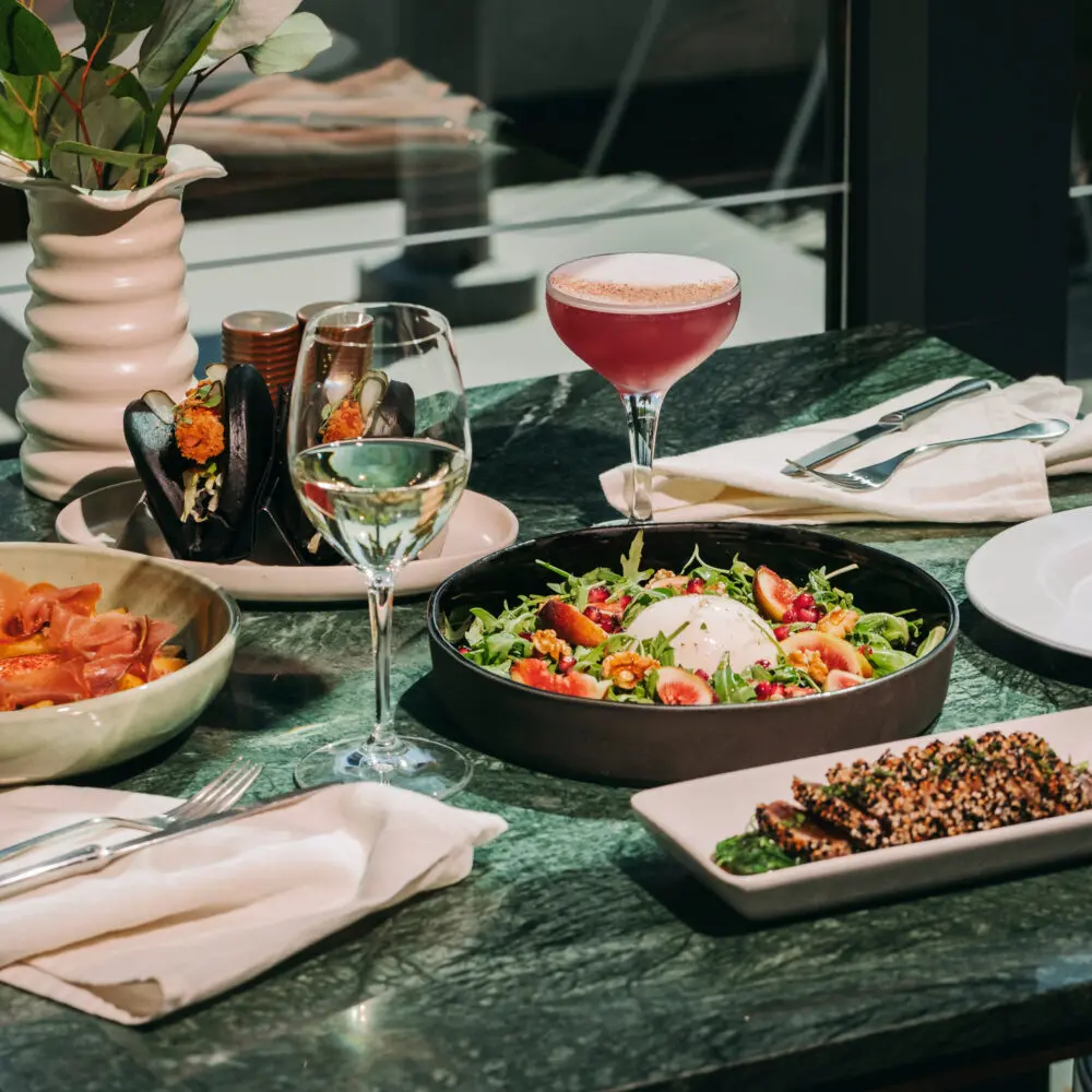 A green marble table set with a salad, mussels, smoked salmon, a pink cocktail, a glass of white wine, and cutlery, with sunlight streaming through a window—evoking the vibrant atmosphere of a rooftop bar in Porto.