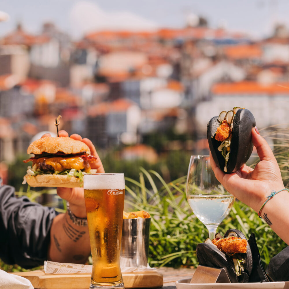 Two people enjoying gourmet burgers and drinks at an outdoor table of a stylish rooftop bar in Porto, with a blurred cityscape featuring red-roofed buildings in the background. One holds a burger, the other a black bun sandwich.