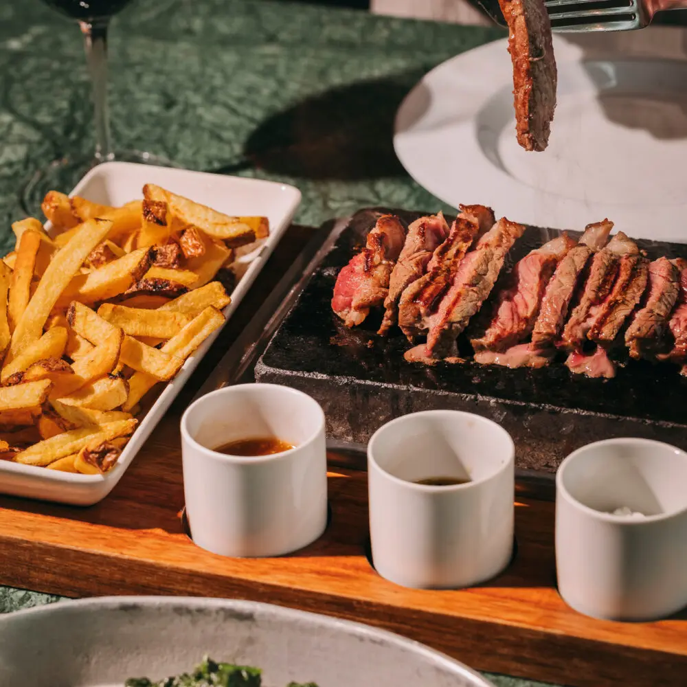 A person cuts into sliced steak on a serving board, accompanied by chips, three sauces, and a side of cooked greens at a vibrant rooftop bar in Porto. A glass of red wine is on the table.