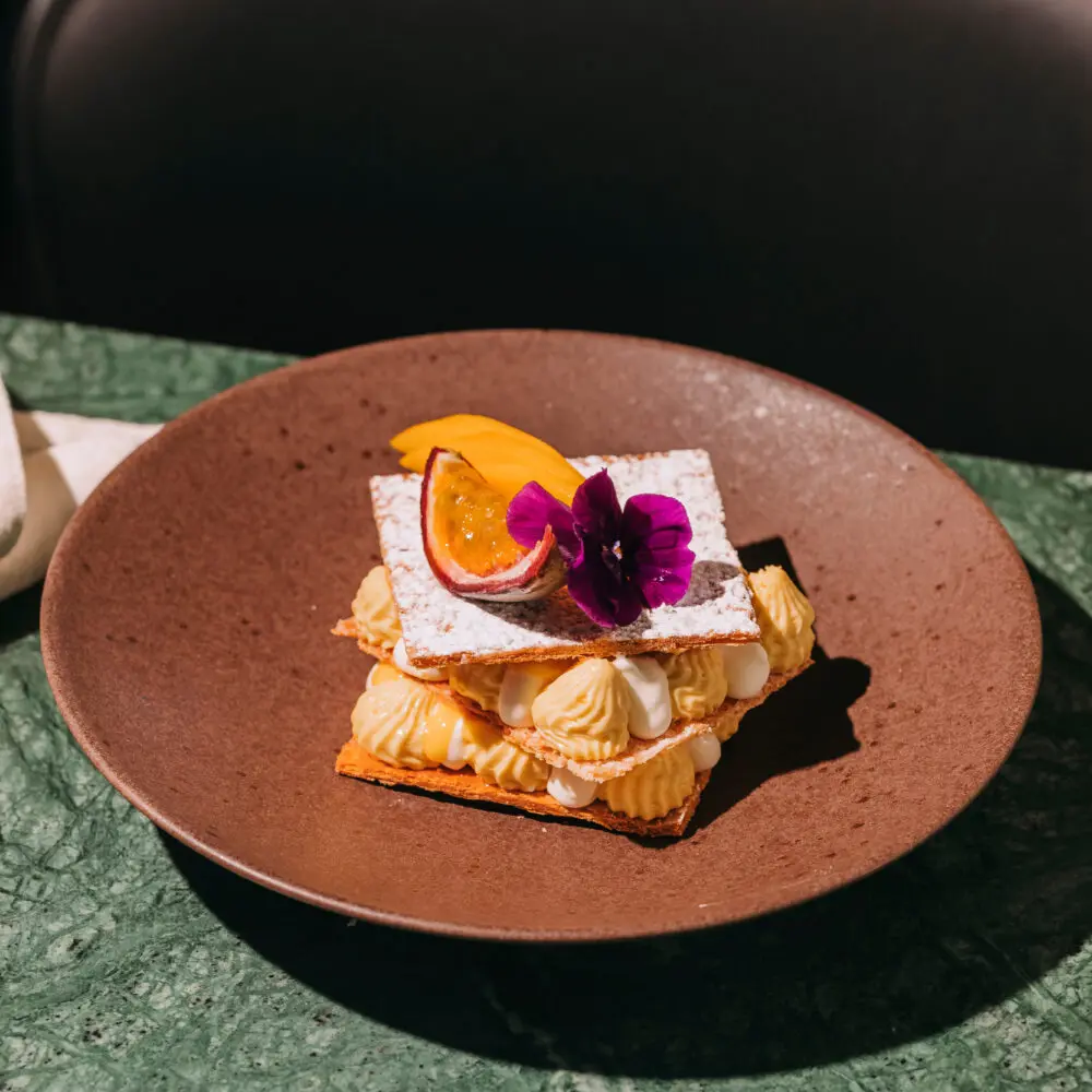 A layered pudding with cream, topped with icing sugar, a slice of fruit, and a purple edible flower, served on a brown plate on a green marble table—perfect for savouring at a stylish rooftop bar in Porto.