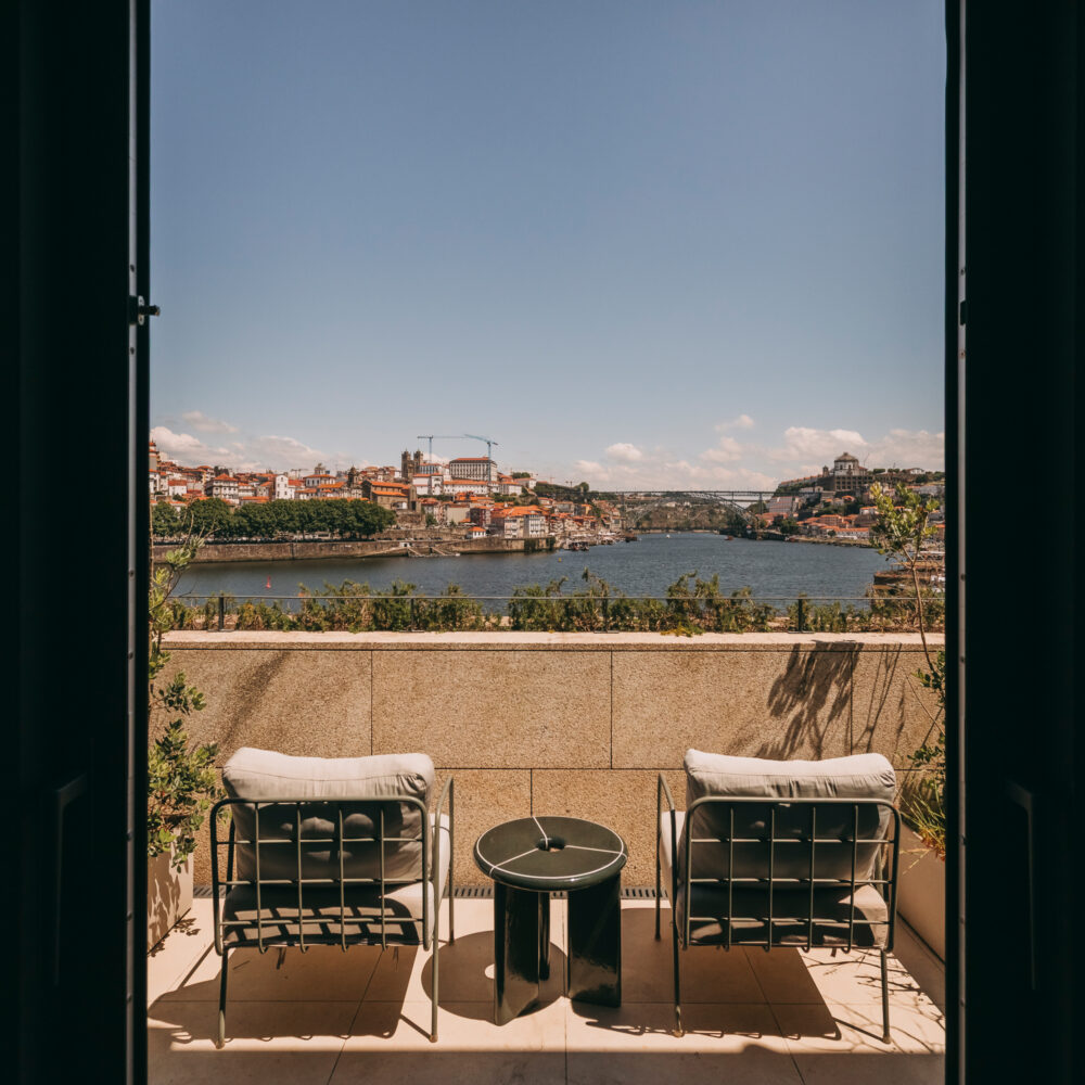 Two chairs and a small table on a patio at Hotel Porto overlook a river and a cityscape under a clear blue sky, framed by an open doorway.