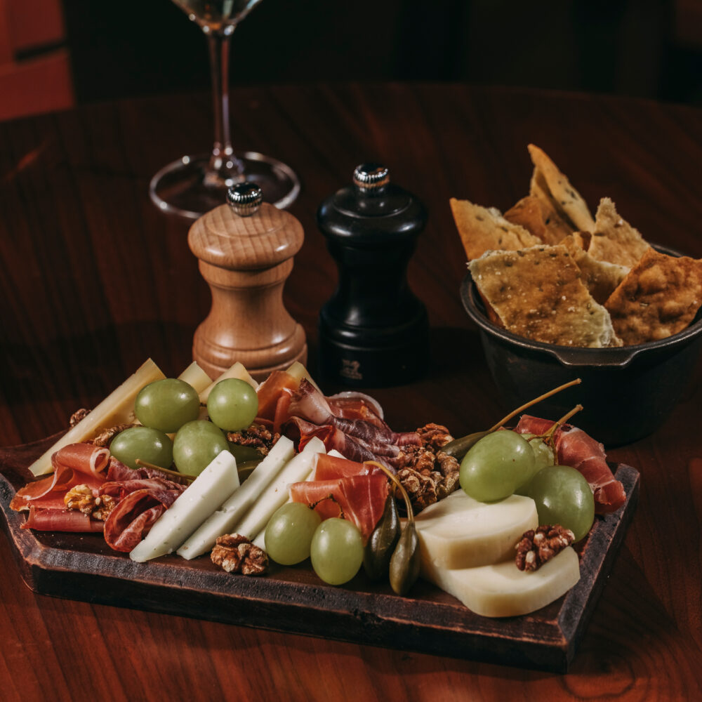 A wooden board with assorted cheeses, prosciutto, green grapes, walnuts, and breadsticks sits on a dark table at a cosy restaurant in Porto, with a bowl of crackers, salt and pepper shakers, and a glass of wine in the background.