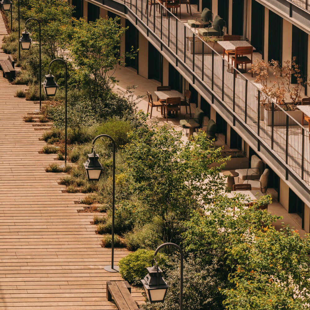 Outdoor terrace with wooden decking, black railing, and potted plants along the walkway. Several tables and chairs are set up on the terrace of this charming hotel in Porto, with greenery and streetlamps lining the area.