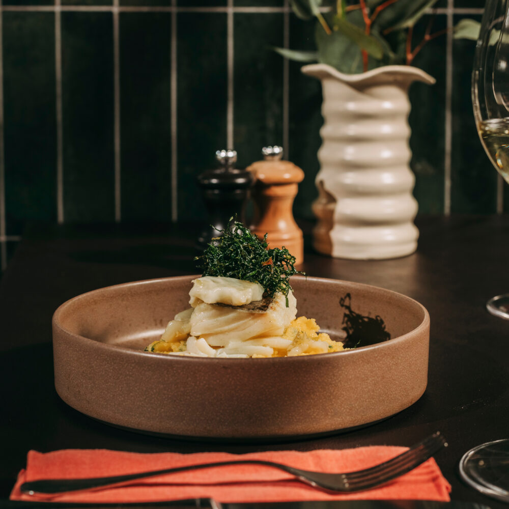 A plated dish with fish and greens sits on a brown plate, showcasing the culinary artistry of a restaurant in Porto. A red serviette with cutlery is in front, and a vase with green leaves along with salt and pepper pots add charm to the background. A wine glass is elegantly positioned to the right.