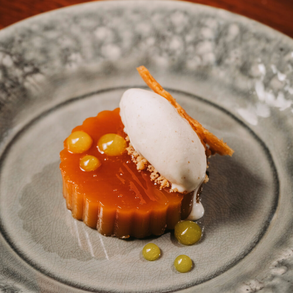 A plated dessert from a renowned restaurant in Porto, featuring a round caramel crème caramel topped with a scoop of vanilla ice cream, a thin crisp, crumbled nuts, and yellow sauce drops, elegantly served on a decorative grey plate.