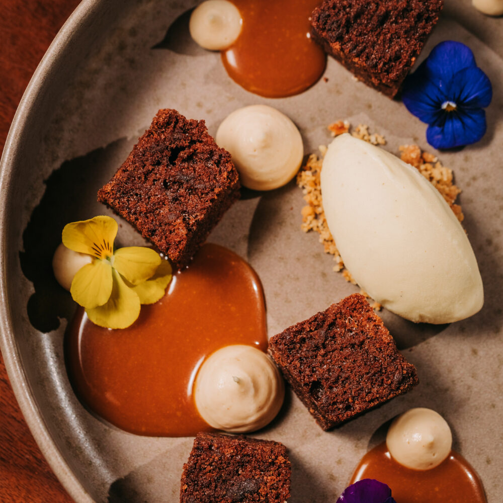 A plate with chocolate cake cubes, caramel sauce, cream dollops, a scoop of ice cream, crumbled topping, and edible yellow and purple flowers is beautifully presented on a brown background—an elegant dessert from a renowned restaurant in Porto.