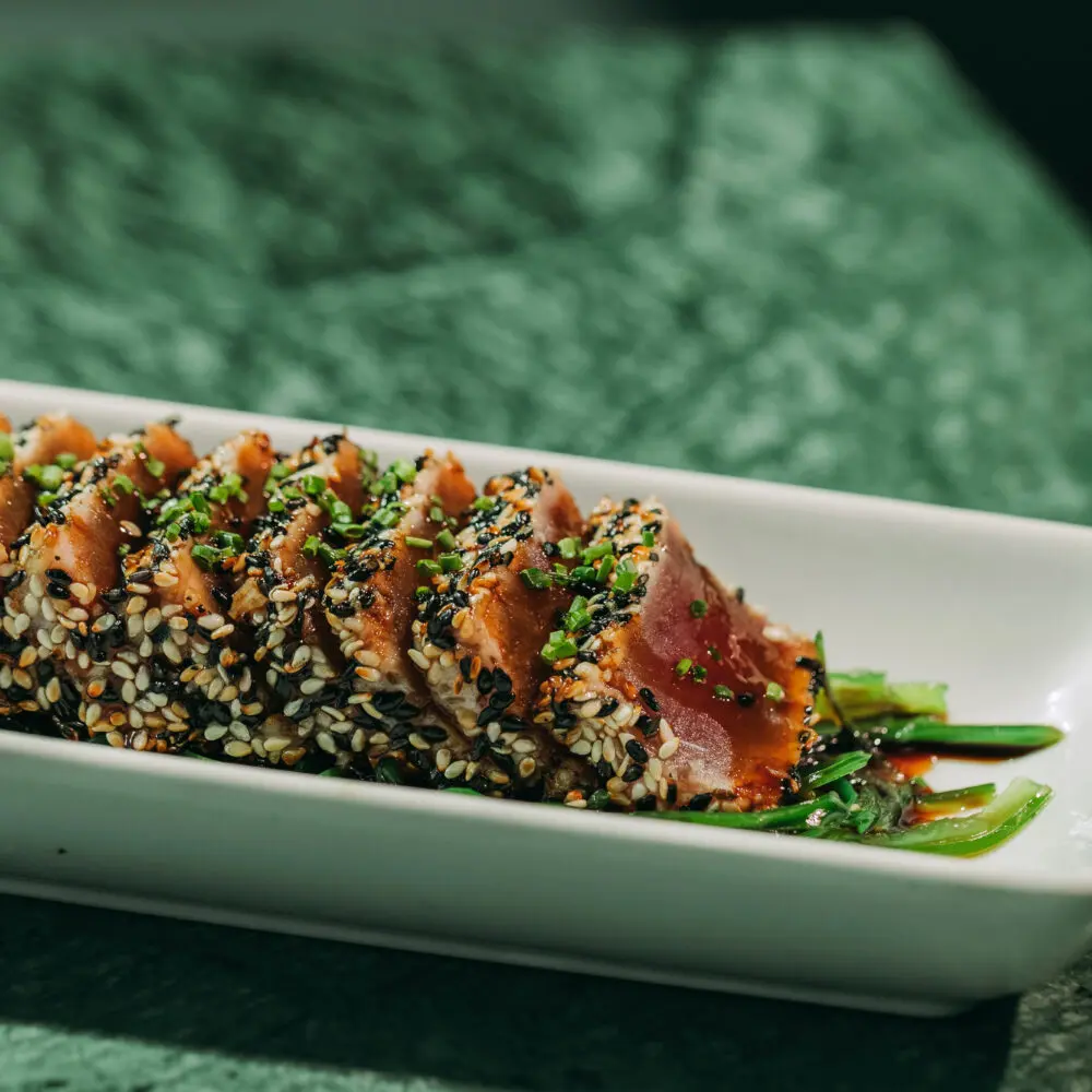 Slices of seared tuna crusted with sesame seeds and garnished with chopped chives, served on a rectangular white plate with a soy-based sauce and green garnish—an elegant dish perfect for enjoying at a rooftop bar in Porto.