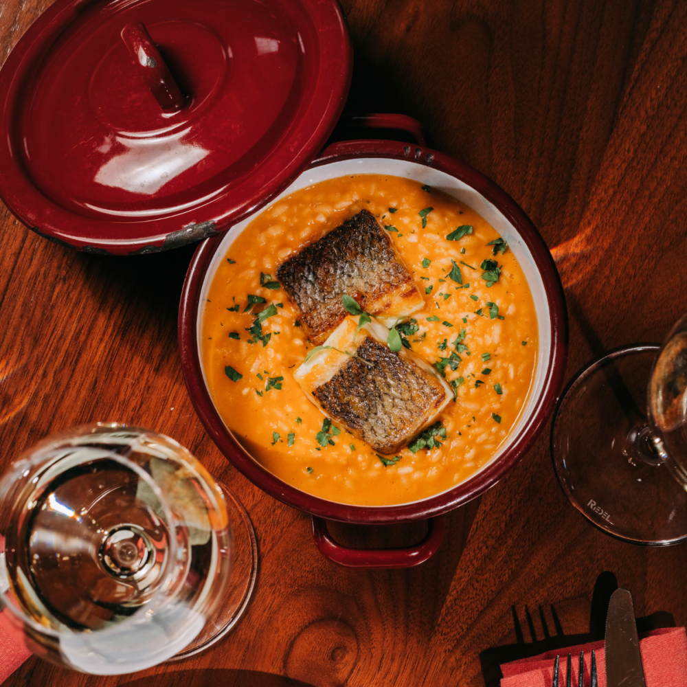 A red pot filled with creamy orange stew, topped with two pieces of grilled fish and chopped herbs, sits on a wooden table next to a glass of white wine and red serviettes with cutlery, capturing the inviting ambience of a restaurant in Porto.