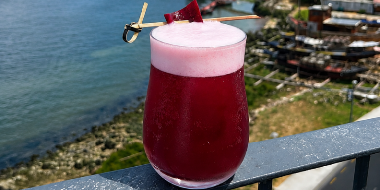 A vibrant purple-red drink with a frothy pink top sits on a balcony rail, garnished with a skewer, overlooking water and boats in the background.