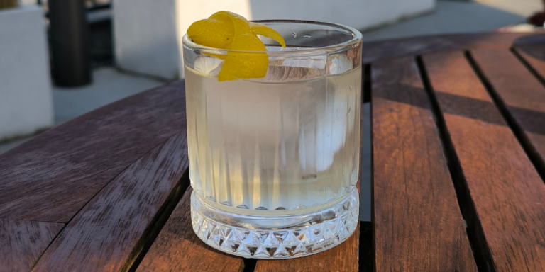 A clear, light-coloured cocktail with a large ice cube, garnished with a bright yellow lemon peel twist. The drink is served in a crystal-cut glass on a dark, slatted wooden table outdoors in warm sunlight.