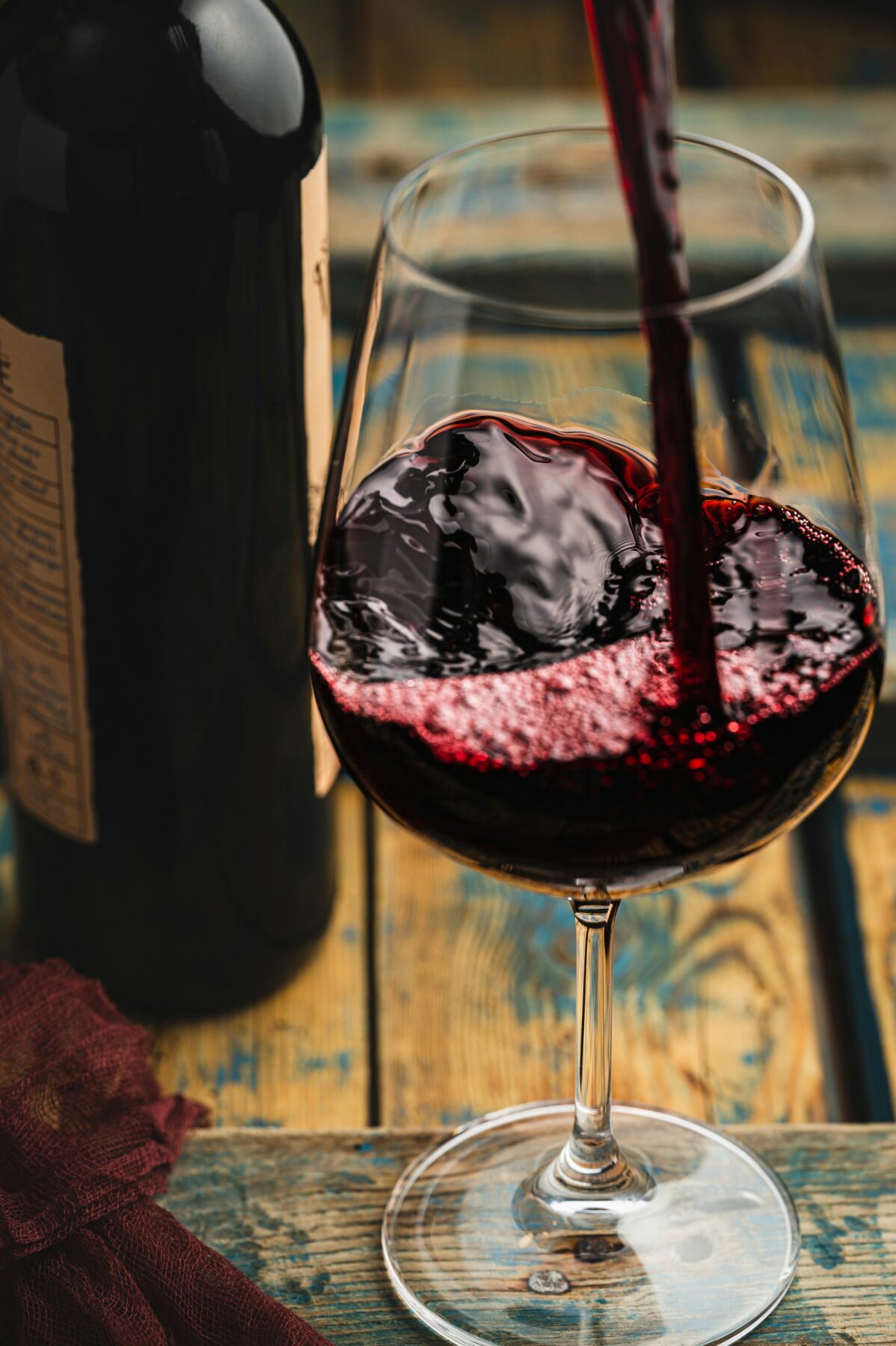 Red wine being poured into a clear wine glass on a rustic wooden table, with a wine bottle and a burgundy cloth nearby.