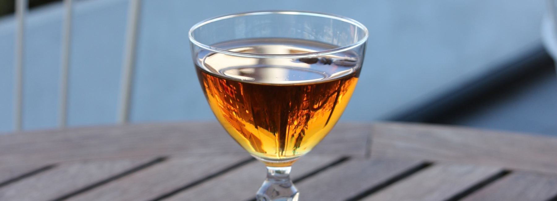 A stemless coupe glass filled with an amber-coloured liquid sits on a slatted wooden outdoor table. Sunlight reflects through the cocktail, creating a warm, golden glow against a soft, blurred blue background.