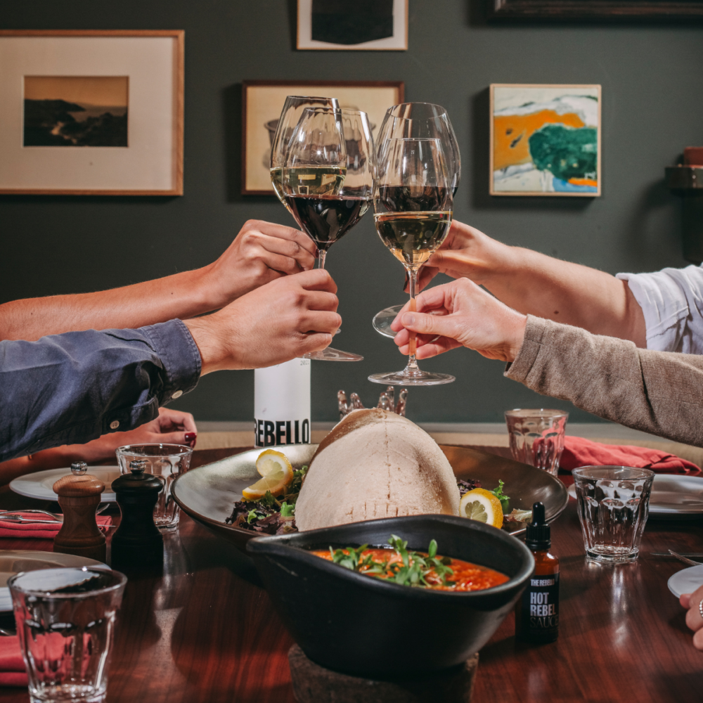 Four people clink wine glasses over a dinner table with a loaf of bread, soup, salad, and condiments—an inviting scene reminiscent of hotel events. Framed art hangs on the dark wall in the background.